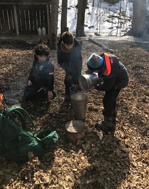 students at The Nature School pour sap into a kettle over a fire to make maple syrup